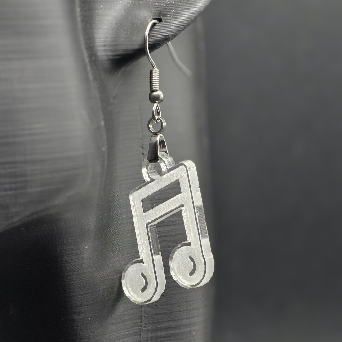 Single earring made of clear acrylic shaped like a beamed music note, hanging from a mannequin ear. Matte black background emphasizes color and shine.