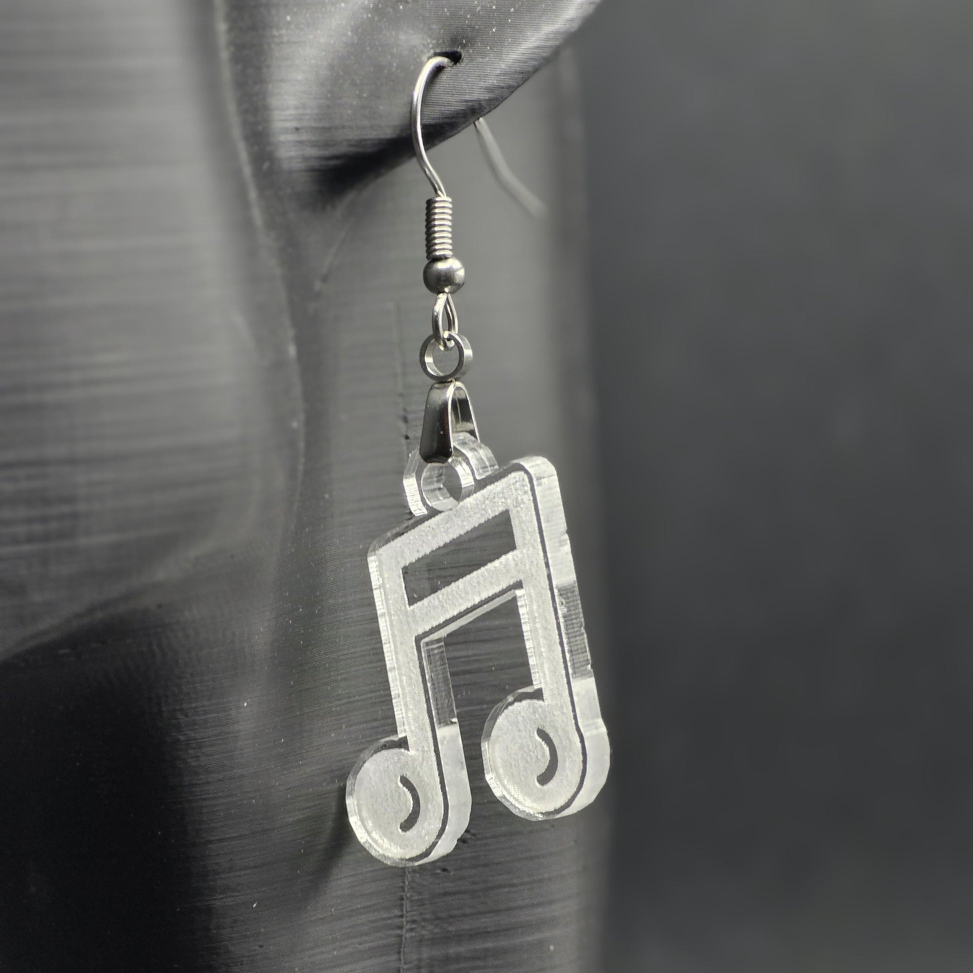 Single earring made of clear acrylic shaped like a beamed music note, hanging from a mannequin ear. Matte black background emphasizes color and shine.