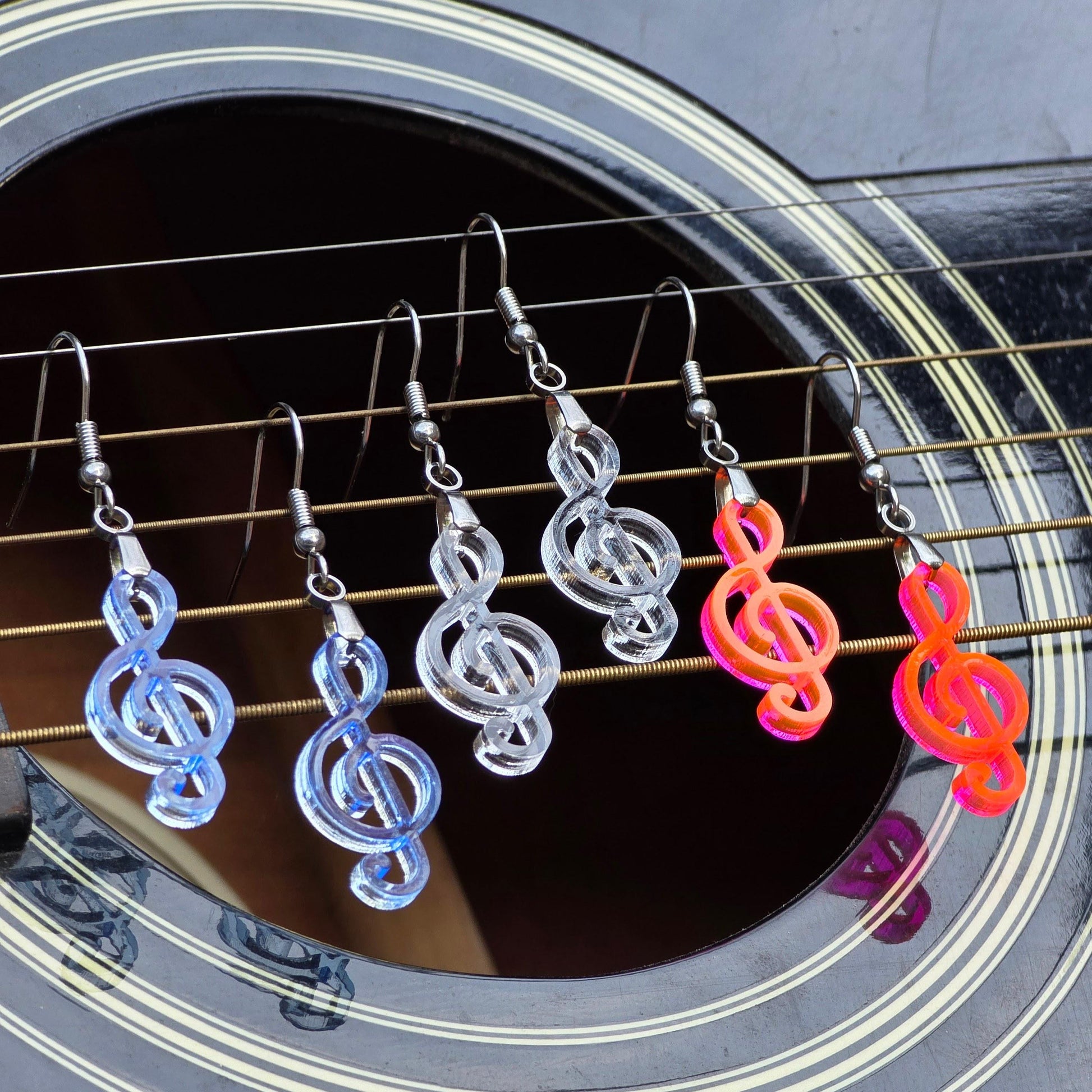 Six handmade treble clef dangle earrings in clear, clear blue, and fluorescent acrylic with stainless steel hooks hang across acoustic guitar strings.