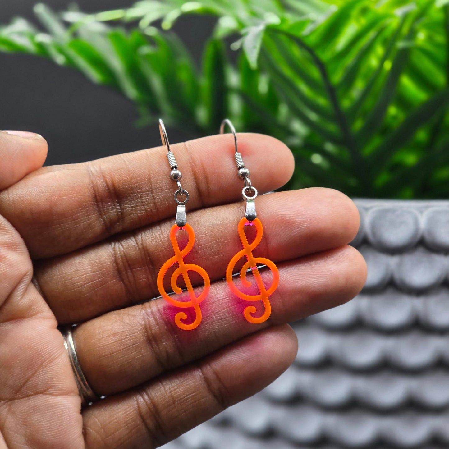 Fluorescent acrylic treble clef earrings with stainless steel hooks held in hand, showing vibrant neon orange-pink color shift. Highlights the lightweight, wearable size of these handmade dangle earrings.