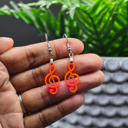 Fluorescent acrylic treble clef earrings with stainless steel hooks held in hand, showing vibrant neon orange-pink color shift. Highlights the lightweight, wearable size of these handmade dangle earrings.