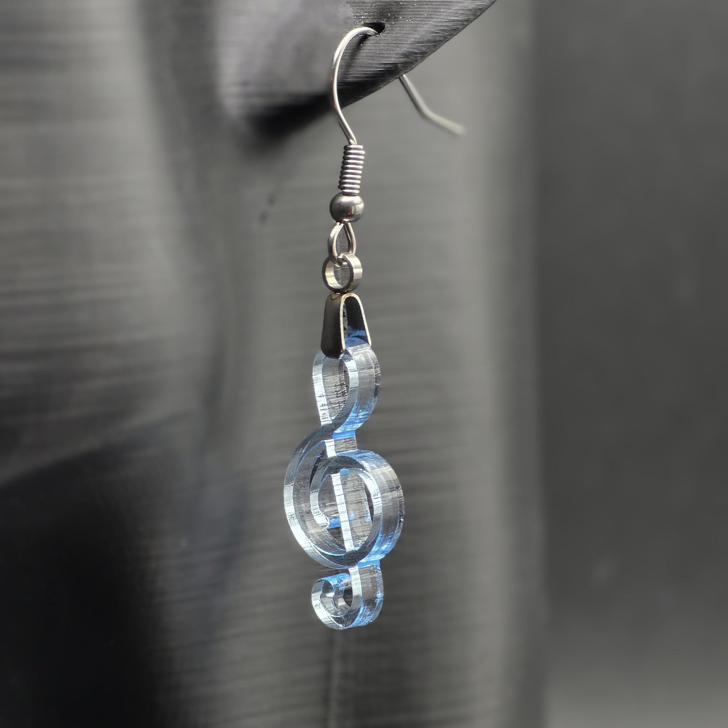 Clear blue acrylic treble clef earring on matte black mannequin ear, showcasing laser-cut detail and stainless steel hardware.