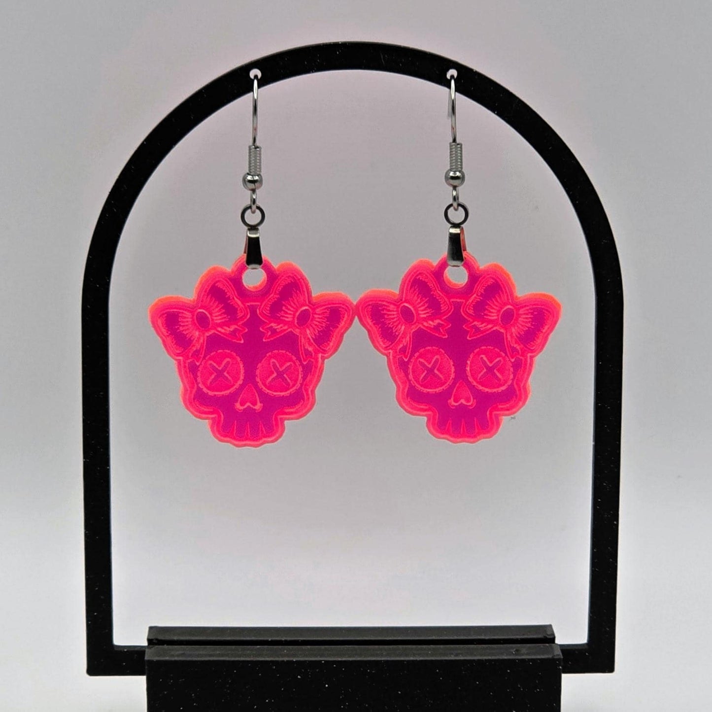 Pair of fluorescent acrylic skull earrings with bows hanging from a black arch-shaped stand. White background enhances the neon glow effect.