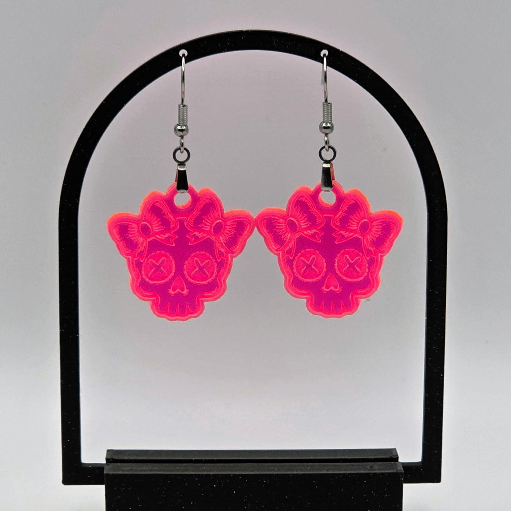 Pair of fluorescent acrylic skull earrings with bows hanging from a black arch-shaped stand. White background enhances the neon glow effect.