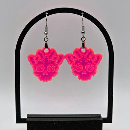 Pair of fluorescent acrylic skull earrings with bows hanging from a black arch-shaped stand. White background enhances the neon glow effect.