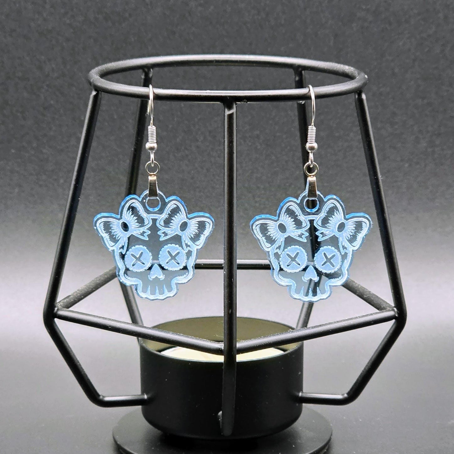 Pair of light blue acrylic skull earrings with bows hanging on a geometric black stand over a black-to-gray gradient background.