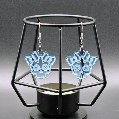 Pair of light blue acrylic skull earrings with bows hanging on a geometric black stand over a black-to-gray gradient background.