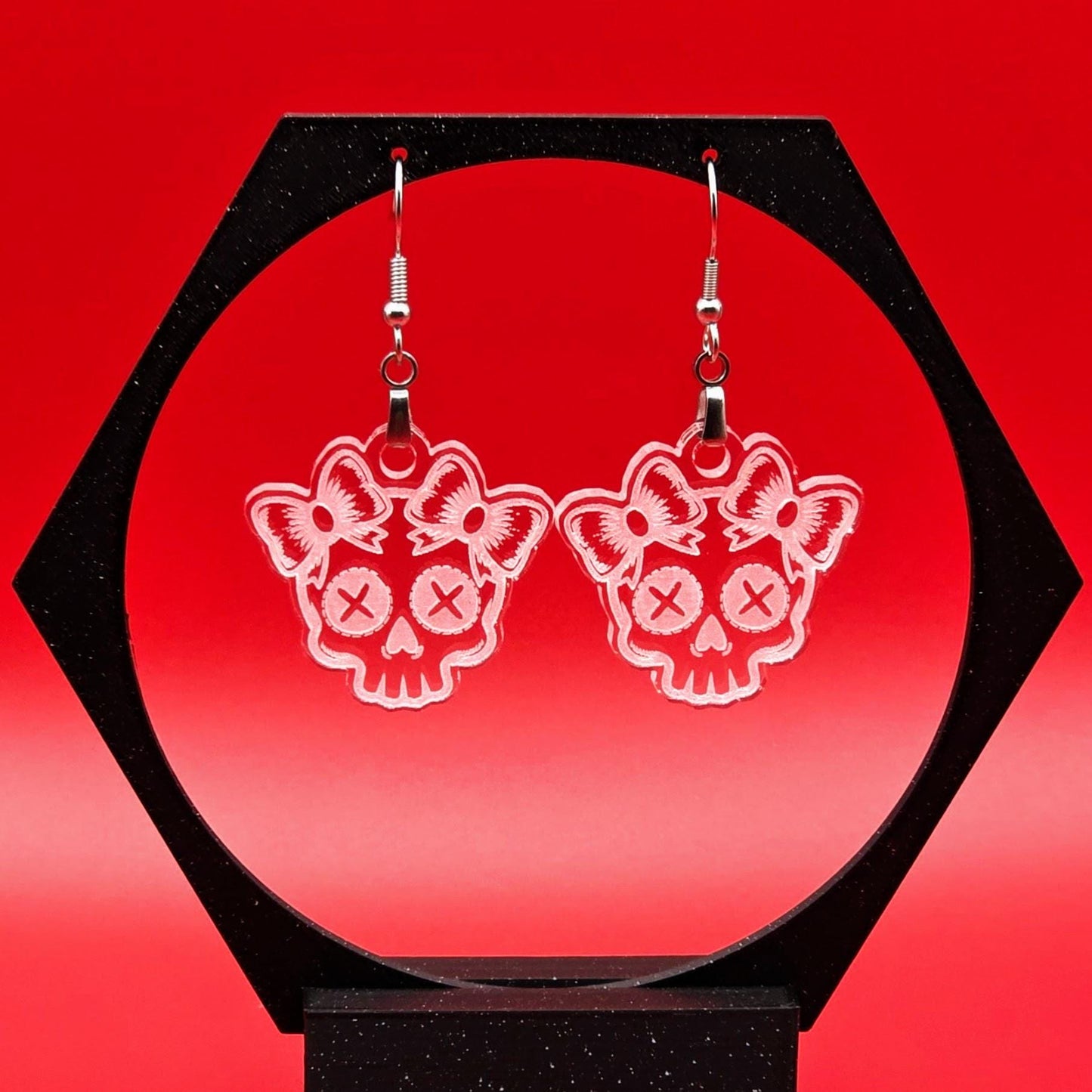 Pair of clear acrylic skull earrings with etched bow designs hanging from a black hexagon display stand. Bright red background adds dramatic contrast.