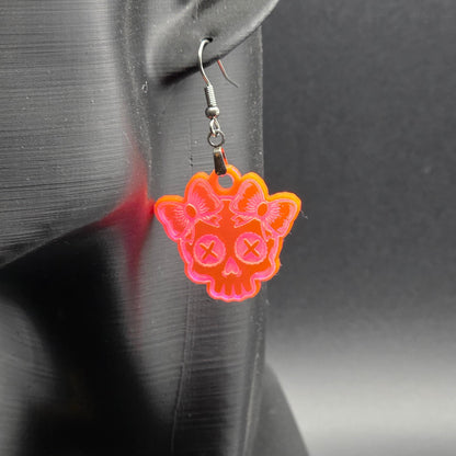 Fluorescent acrylic skull earring with bow accents displayed on a black mannequin ear. Dark background emphasizes vivid neon color.