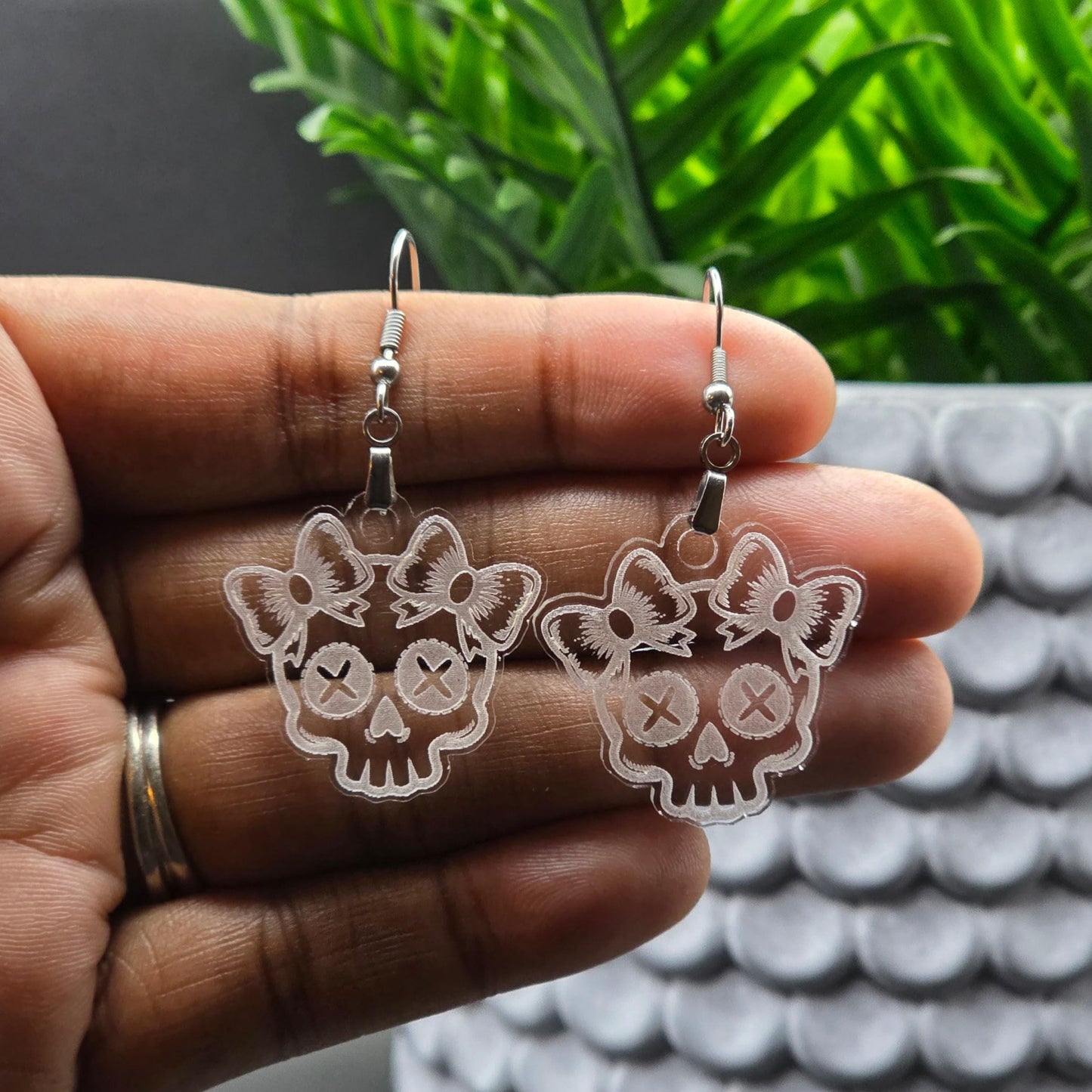 Pair of clear acrylic skull earrings with bows displayed on an open palm in natural light. Green plant and textured white planter in the background.