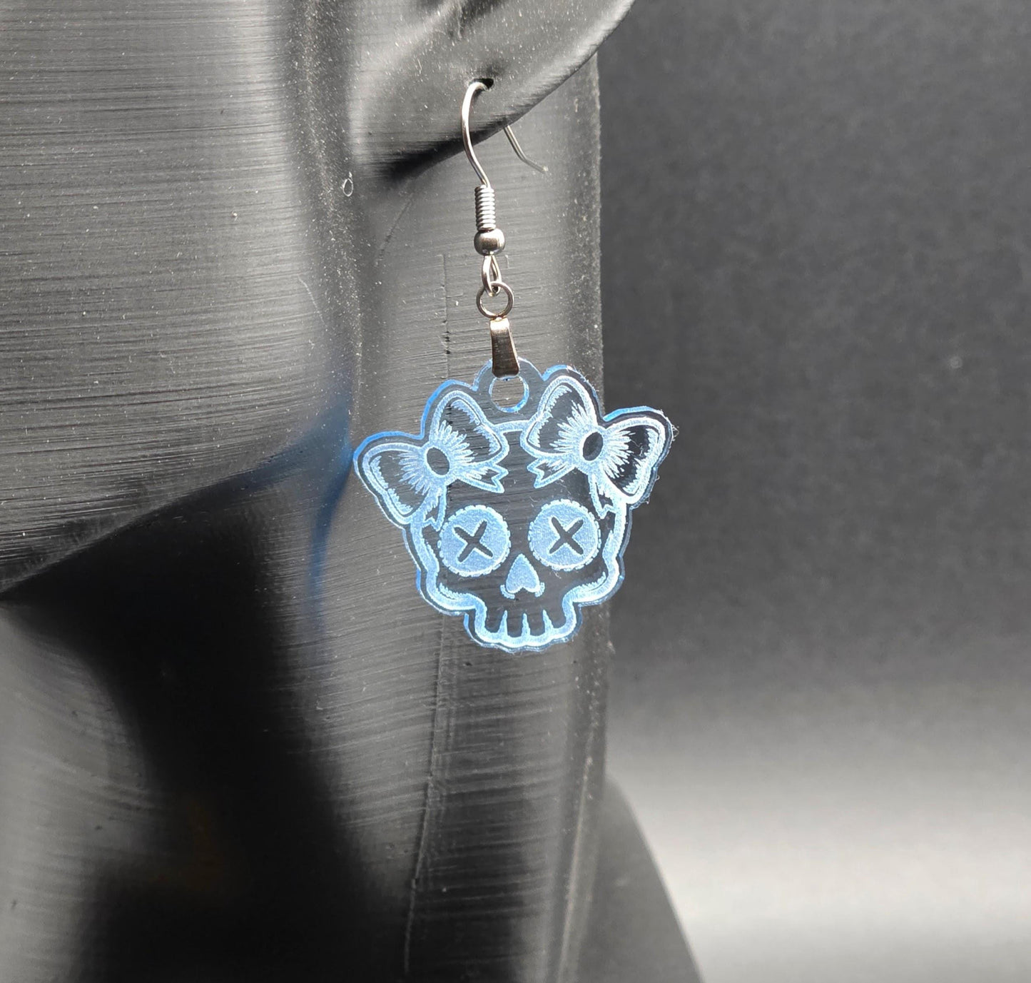 Clear blue acrylic skull earring with etched bows displayed on a black mannequin ear against a neutral gray background.