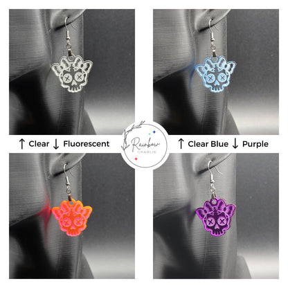 Four skull-shaped acrylic earrings with bows displayed on a mannequin. Top left: clear; top right: clear blue; bottom left: fluorescent; bottom right: purple. Central Rainbow Charlie logo with labeled color directions.