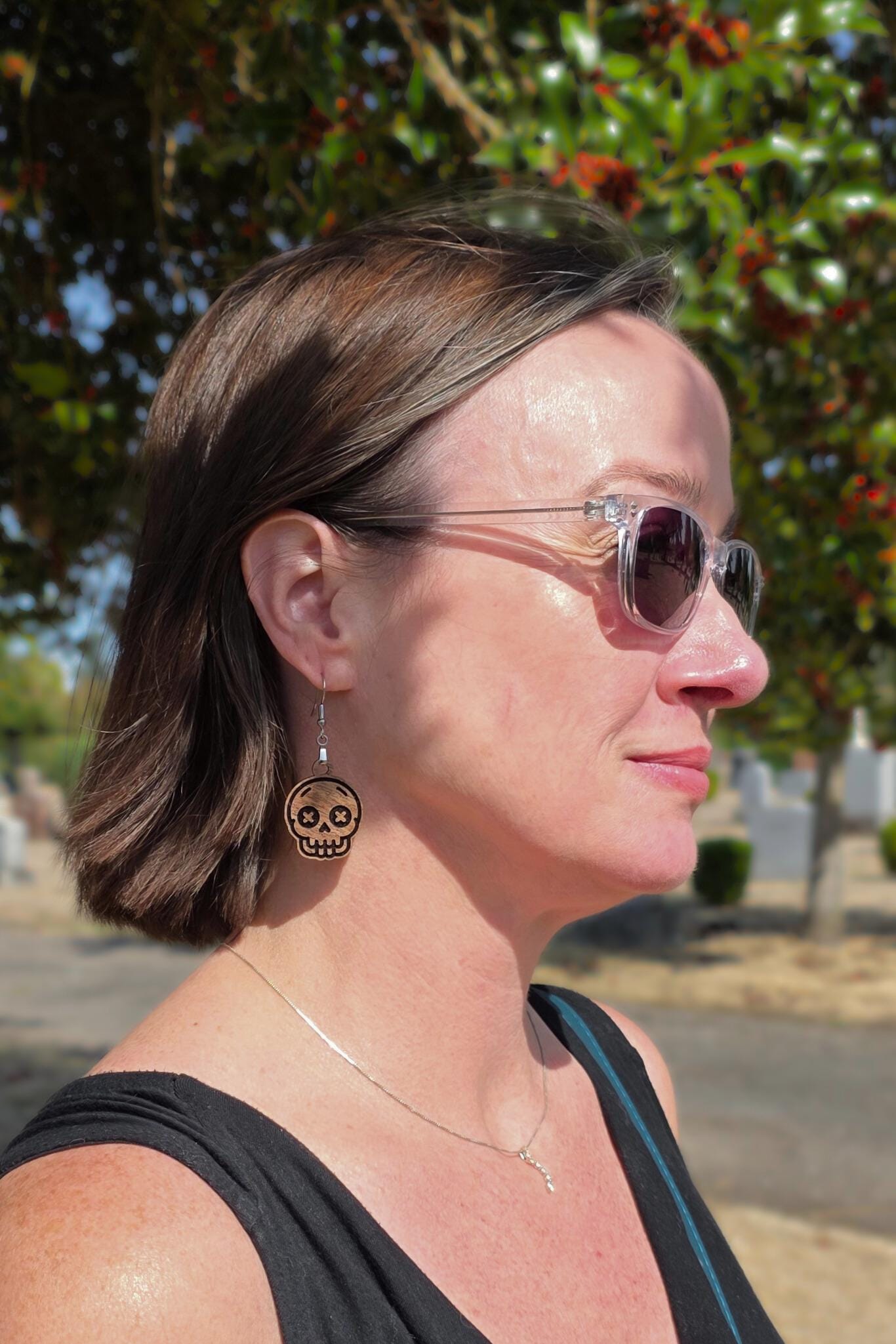 Outdoor close-up of a woman wearing wooden skull earrings with X-shaped eyes and engraved details. She’s standing under a tree with red berries, wearing sunglasses and a black top. Earrings dangle gently from her ears, showcasing their lightweight, wearable design.