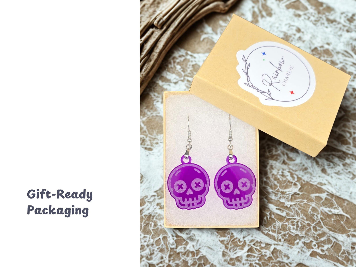 A pair of purple skull earrings displayed in a kraft gift box with soft white padding. The box lid features the Rainbow Charlie logo sticker. Set atop a marbled surface, the image includes the phrase “Gift-Ready Packaging” on the left, emphasizing presentation quality.