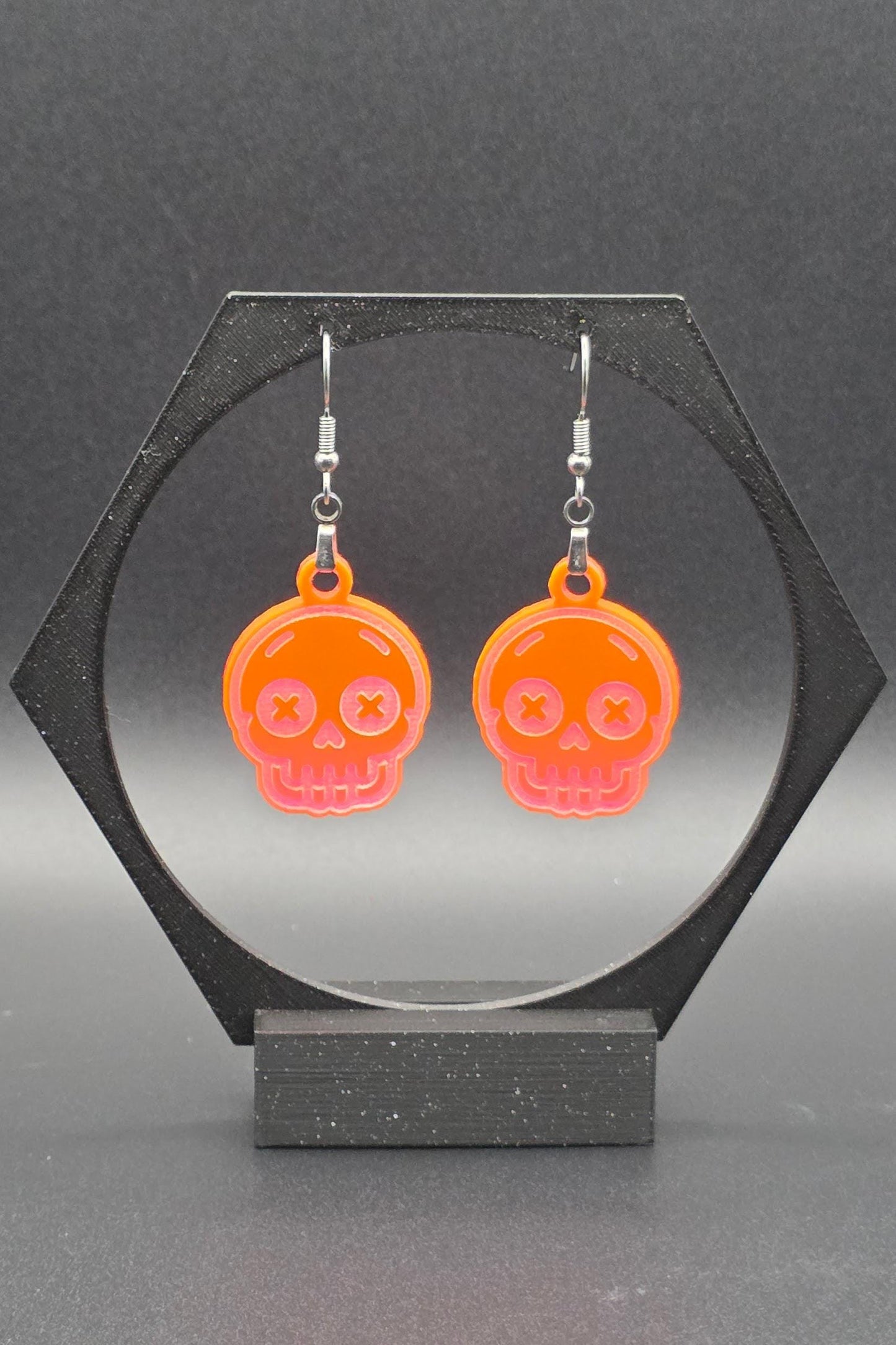 A pair of fluorescent acrylic skull earrings with laser-engraved features, including X-shaped eyes and a toothy grin, displayed on a geometric black stand against a gray background.