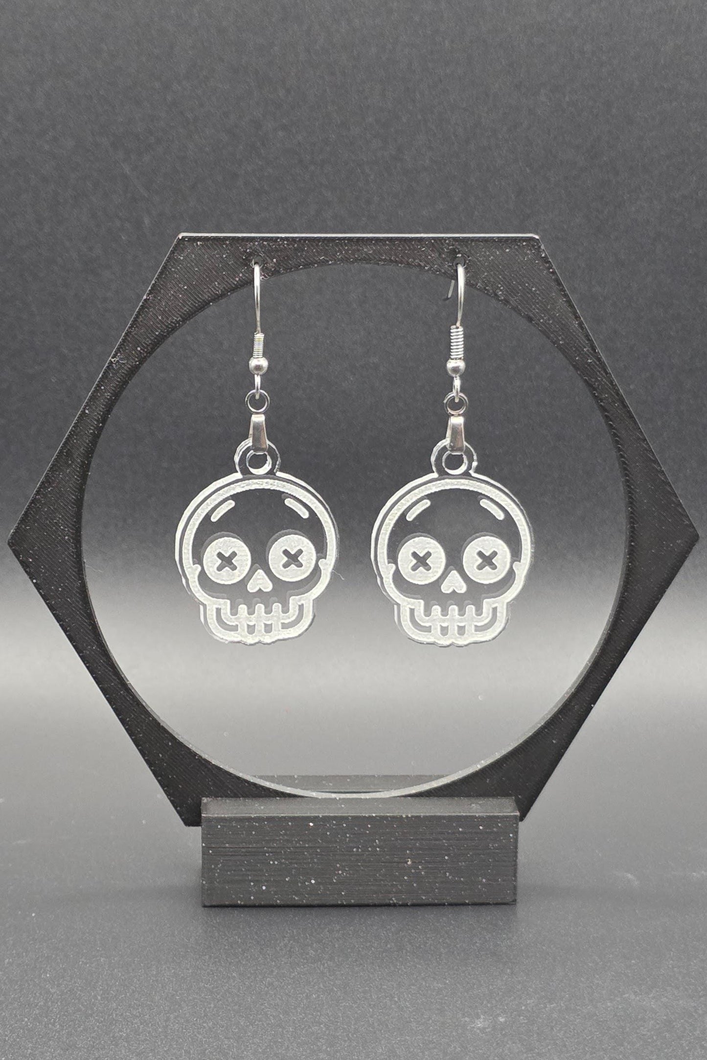 A pair of clear acrylic skull earrings with laser-engraved features, including X-shaped eyes and a toothy grin, displayed on a geometric black stand against a gray background.