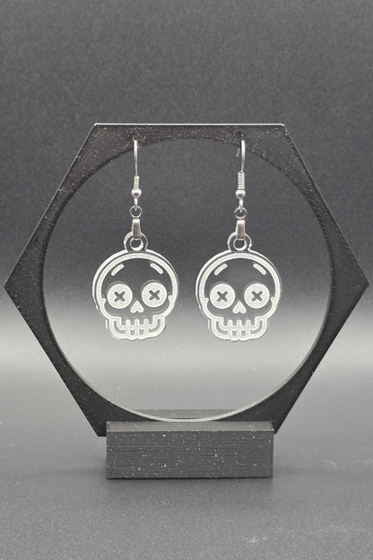 A pair of clear acrylic skull earrings with laser-engraved features, including X-shaped eyes and a toothy grin, displayed on a geometric black stand against a gray background.