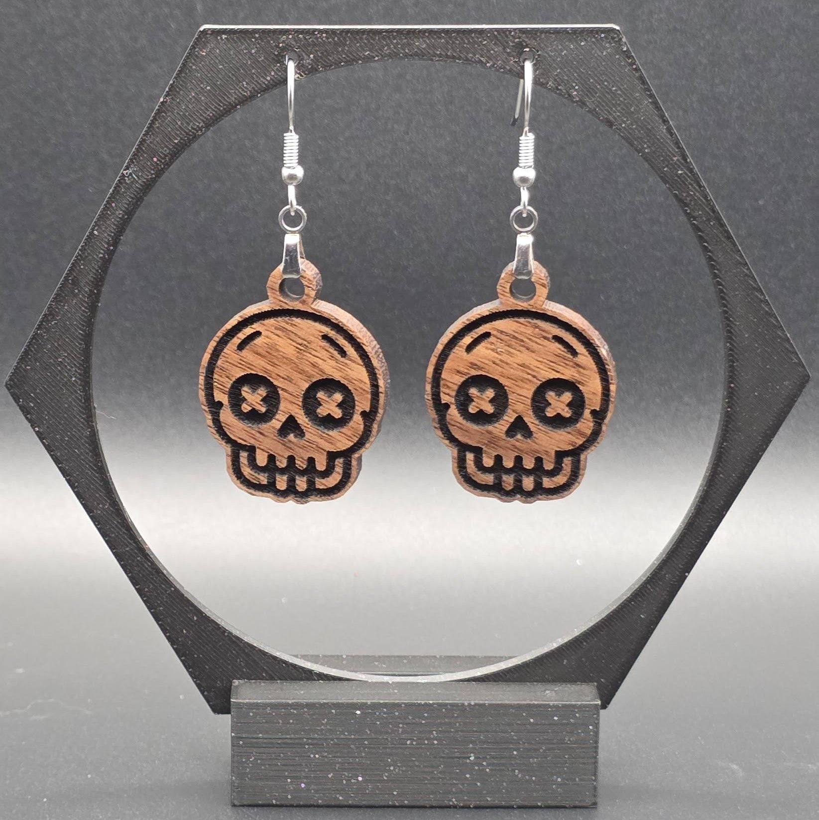 A pair of walnut hardwood skull earrings with laser-engraved features, including X-shaped eyes and a toothy grin, displayed on a geometric black stand against a gray background.