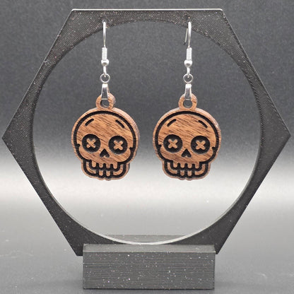 A pair of walnut hardwood skull earrings with laser-engraved features, including X-shaped eyes and a toothy grin, displayed on a geometric black stand against a gray background.