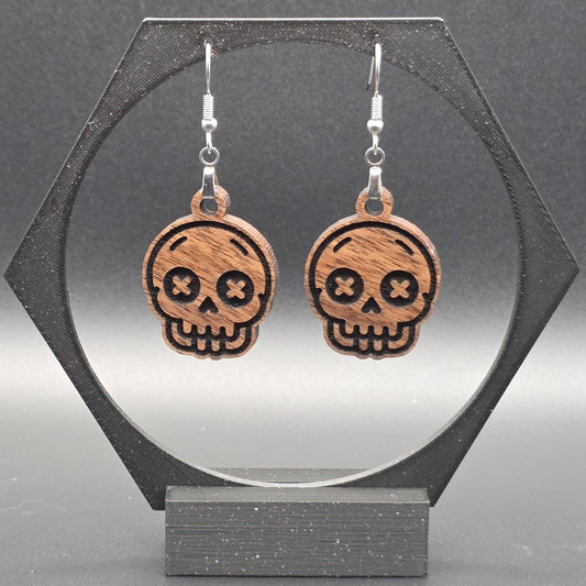 A pair of walnut hardwood skull earrings with laser-engraved features, including X-shaped eyes and a toothy grin, displayed on a geometric black stand against a gray background.