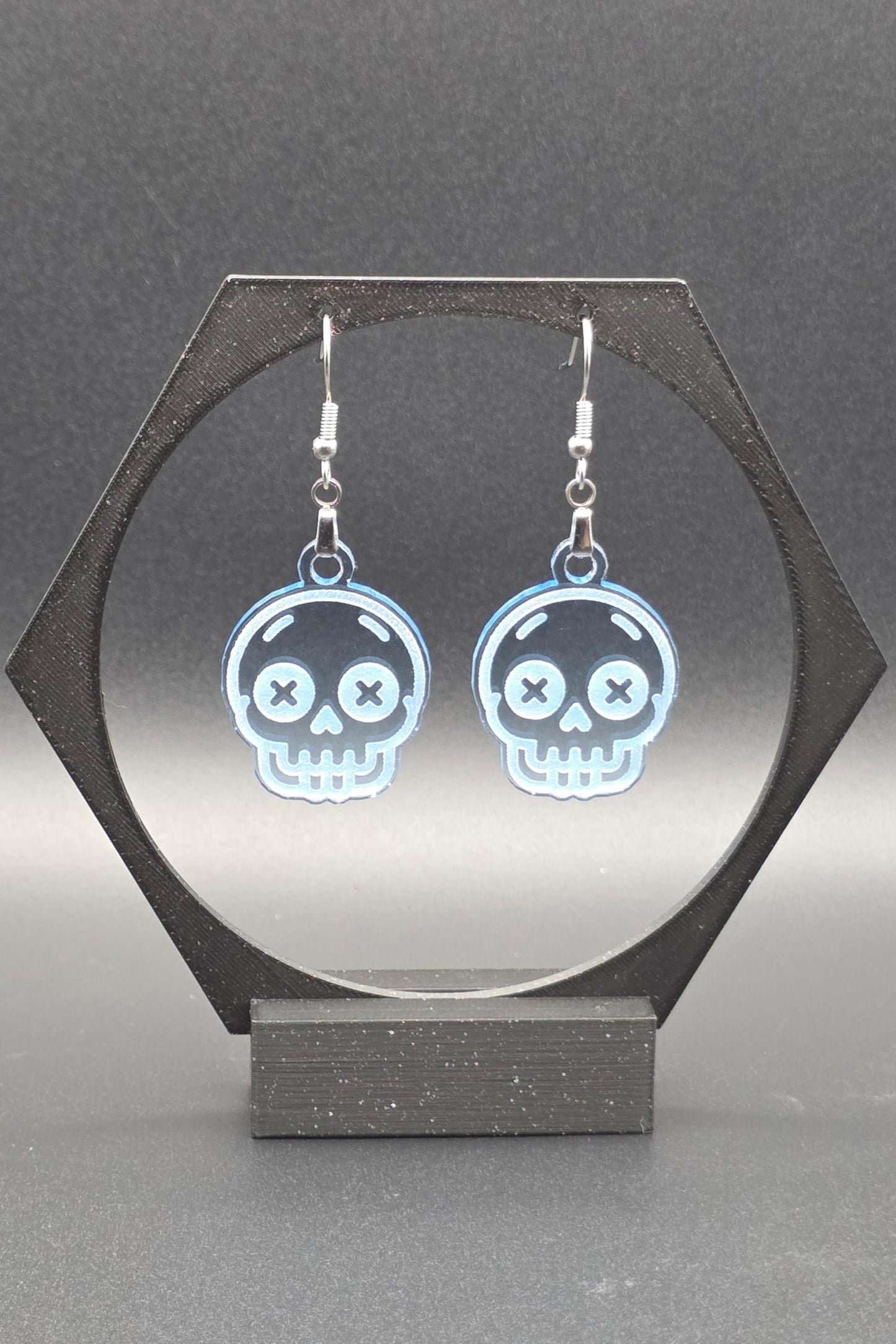 A pair of translucent blue acrylic skull earrings with laser-engraved features, including X-shaped eyes and a toothy grin, displayed on a geometric black stand against a gray background.