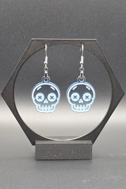 A pair of translucent blue acrylic skull earrings with laser-engraved features, including X-shaped eyes and a toothy grin, displayed on a geometric black stand against a gray background.