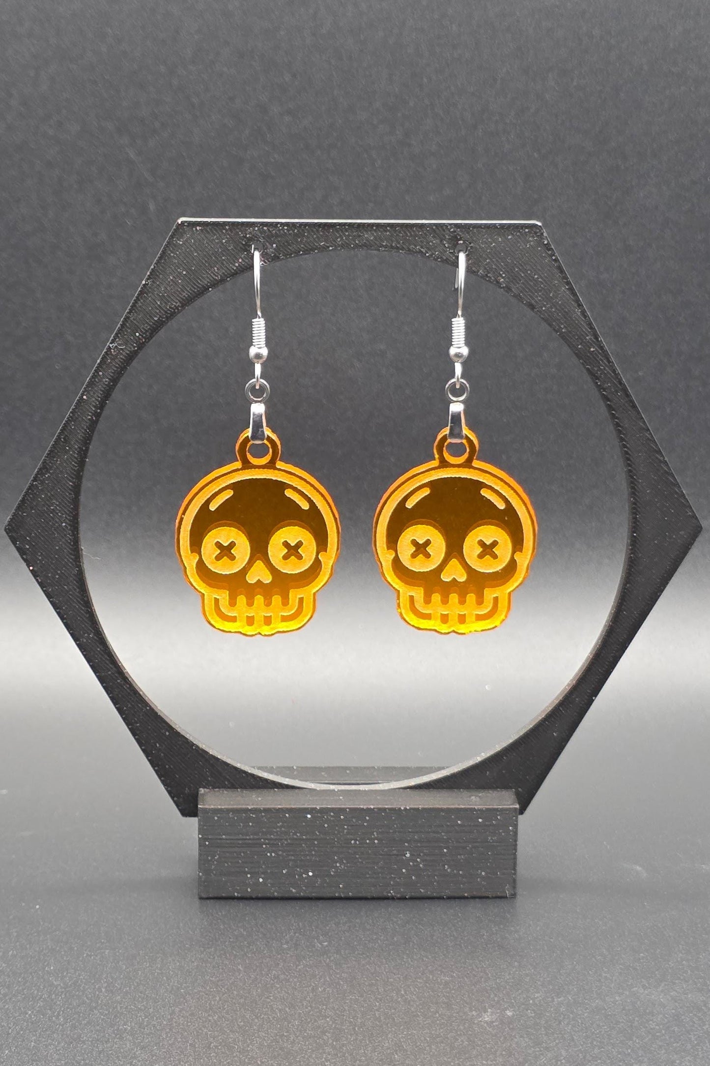 A pair of orange acrylic skull earrings with laser-engraved features, including X-shaped eyes and a toothy grin, displayed on a geometric black stand against a gray background.