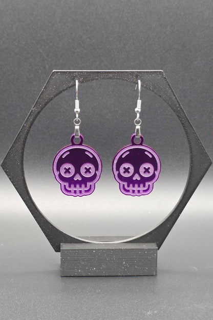 A pair of purple acrylic skull earrings with laser-engraved features, including X-shaped eyes and a toothy grin, displayed on a geometric black stand against a gray background.