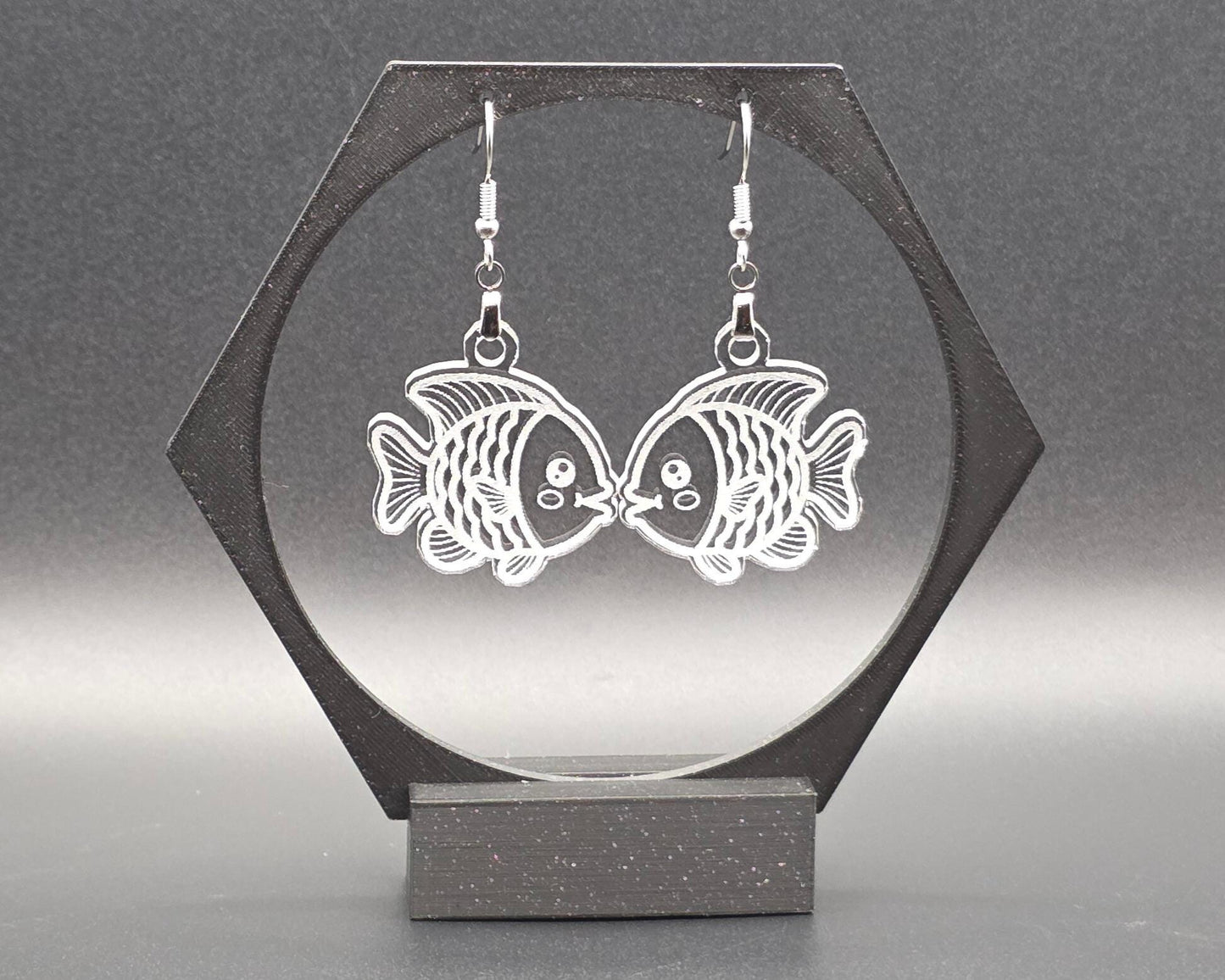 A pair of clear laser-cut acrylic goldfish earrings with nickel-free hooks displayed on a black hexagon earring stand. The transparent fish design shows fine engraved details, creating a whimsical, aquatic jewelry look.