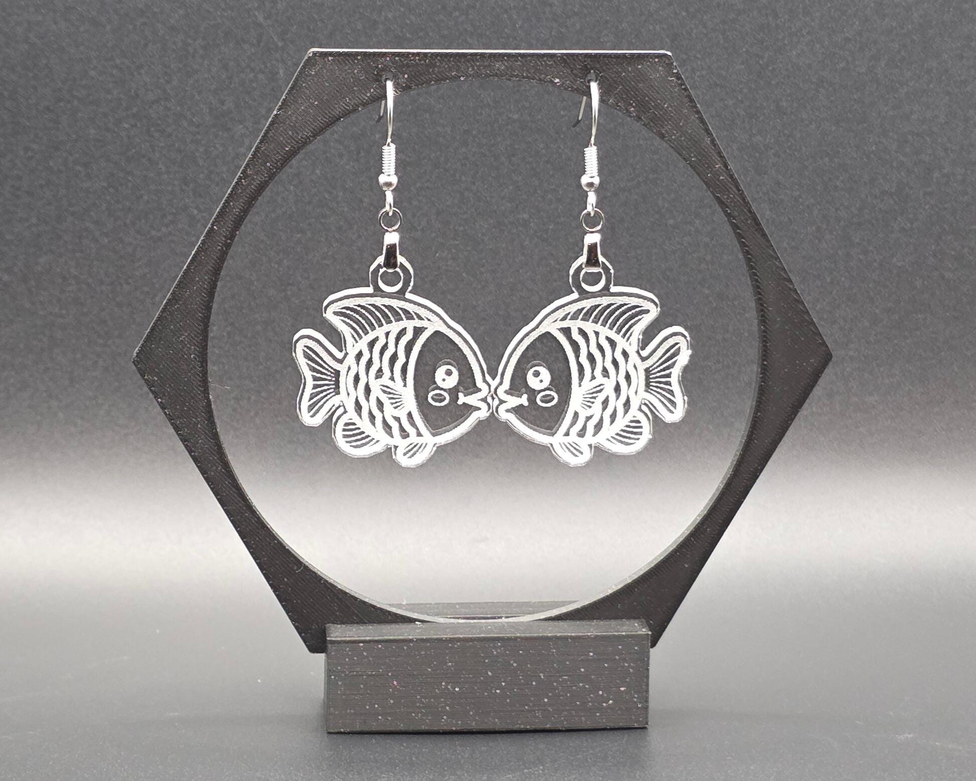 A pair of clear laser-cut acrylic goldfish earrings with nickel-free hooks displayed on a black hexagon earring stand. The transparent fish design shows fine engraved details, creating a whimsical, aquatic jewelry look.