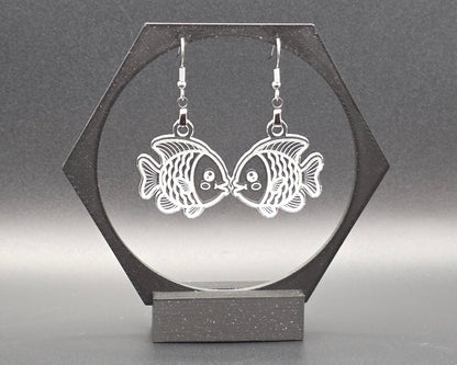 A pair of clear laser-cut acrylic goldfish earrings with nickel-free hooks displayed on a black hexagon earring stand. The transparent fish design shows fine engraved details, creating a whimsical, aquatic jewelry look.