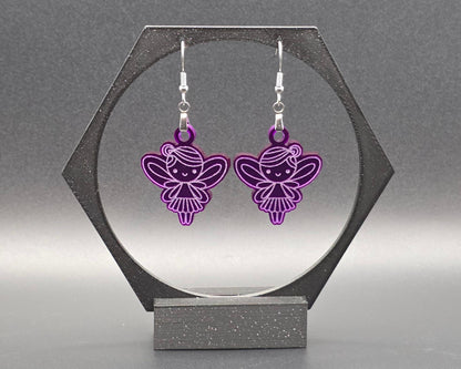 A pair of purple acrylic fairy earrings with engraved wing and dress details, hanging from nickel-free stainless steel hooks, displayed in a black hexagonal jewelry stand against a soft gray background. Handmade by Rainbow Charlie, whimsical and lightweight.