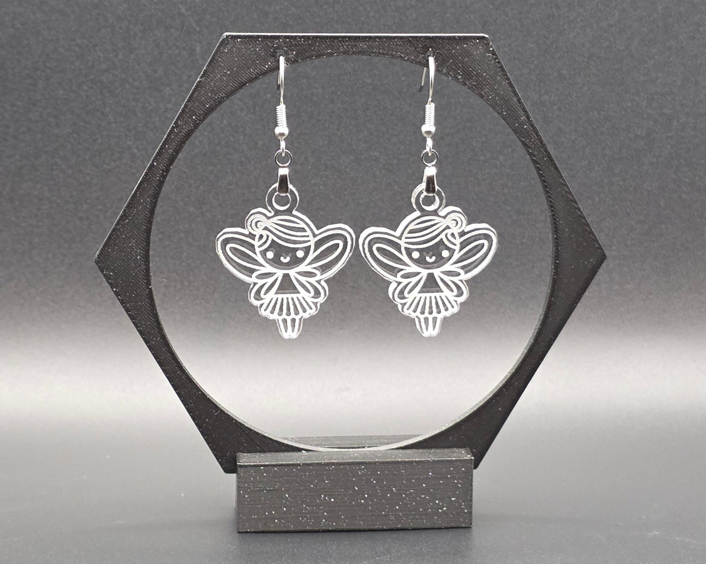 A pair of clear acrylic fairy earrings with engraved wing and dress details, hanging from nickel-free stainless steel hooks, displayed in a black hexagonal jewelry stand against a soft gray background. Handmade by Rainbow Charlie, whimsical and lightweight.