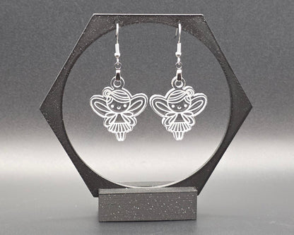 A pair of clear acrylic fairy earrings with engraved wing and dress details, hanging from nickel-free stainless steel hooks, displayed in a black hexagonal jewelry stand against a soft gray background. Handmade by Rainbow Charlie, whimsical and lightweight.