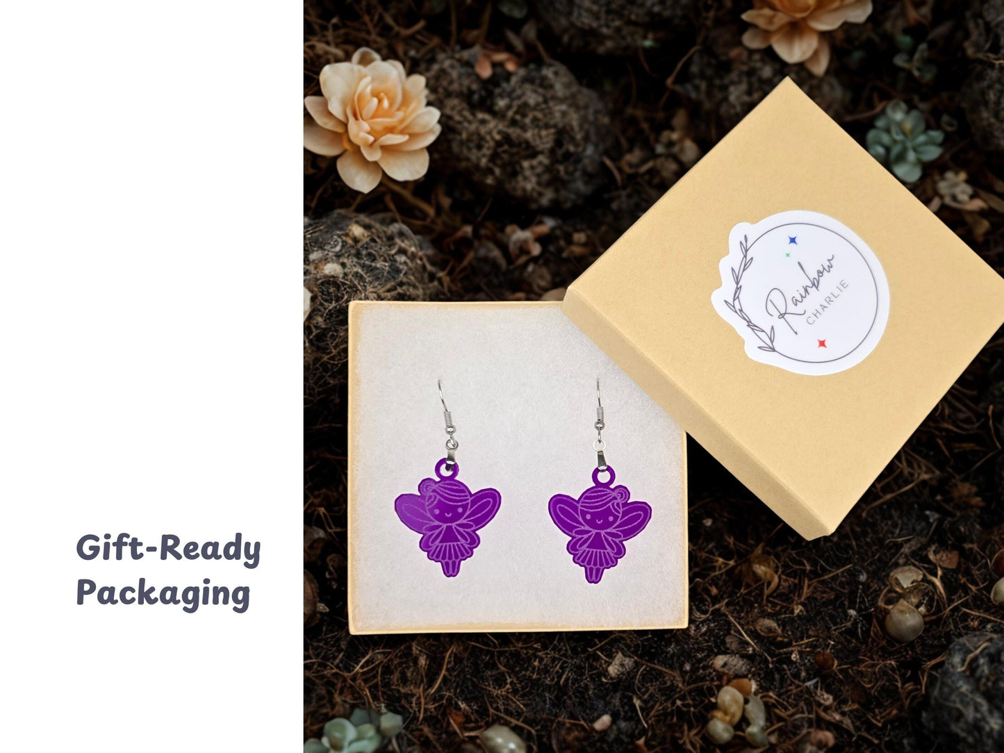 Pair of purple acrylic fairy earrings displayed in a beige jewelry gift box with the Rainbow Charlie logo on the lid. Surrounded by soft-focus succulents, the image highlights the handmade earrings’ ready-to-gift packaging.