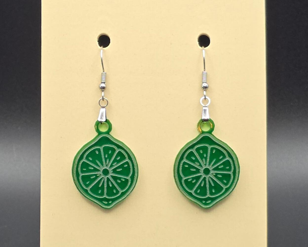 A pair of green acrylic lime slice earrings with stainless steel hooks hanging on a beige display card. The earrings are translucent and etched with citrus segment details for a fresh, juicy look.