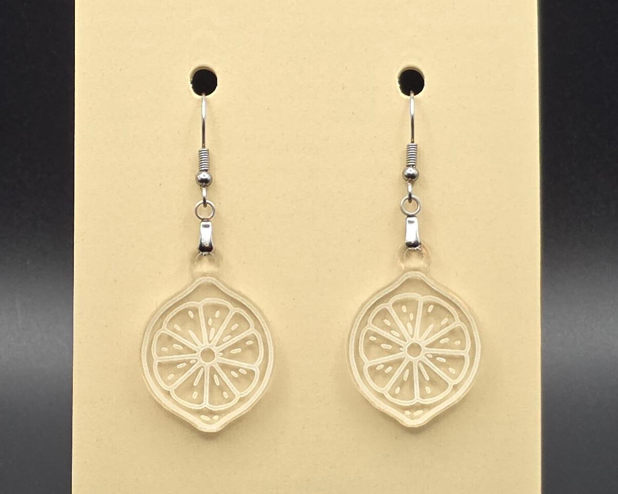 A pair of clear acrylic citrus slice earrings on a beige earring card. The transparent design shows engraved fruit segment lines, creating a subtle, minimalist look.