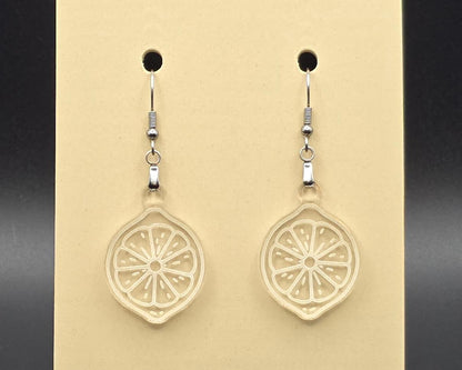 A pair of clear acrylic citrus slice earrings on a beige earring card. The transparent design shows engraved fruit segment lines, creating a subtle, minimalist look.