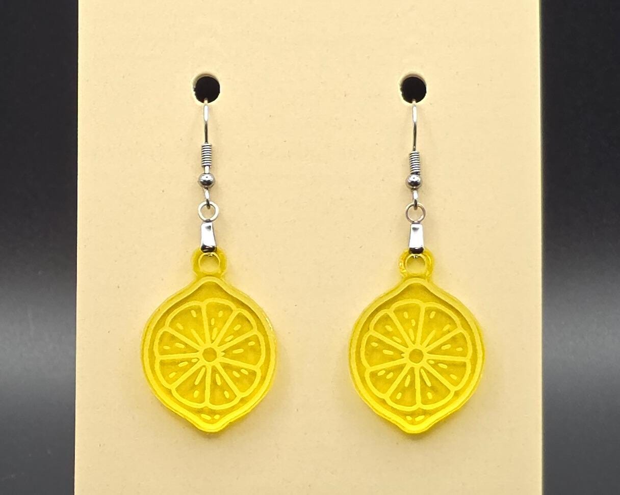 A pair of yellow acrylic lemon slice earrings with stainless steel hooks displayed on a beige earring card. The citrus slices are bright, translucent, and laser-cut with detailed segment etching.