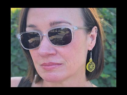 A woman wearing clear sunglasses and lemon yellow acrylic citrus slice earrings outdoors. The translucent earrings catch the sunlight, showing detailed laser-cut fruit segments.