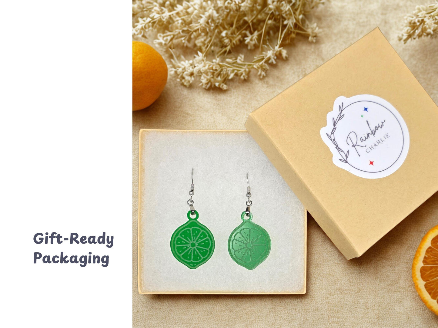 A pair of green acrylic lime slice earrings displayed inside a beige gift box lined with white padding. The box lid, featuring a Rainbow Charlie sticker, is set beside the earrings with citrus fruit and dried flowers around it.