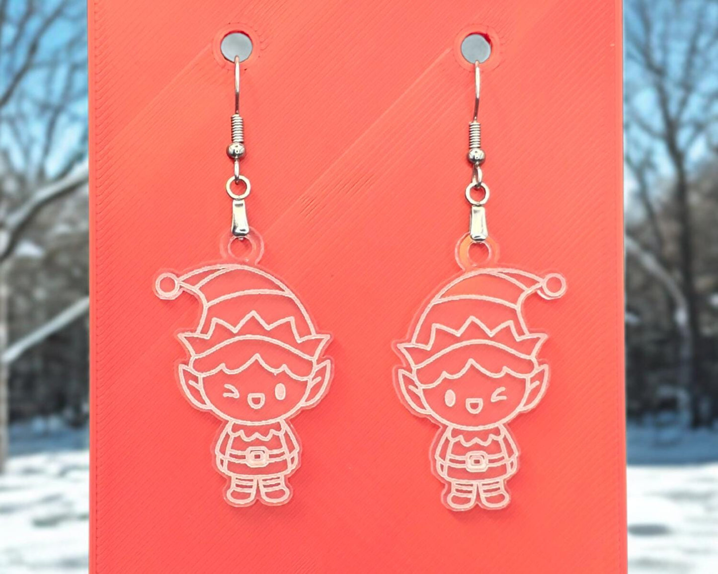 Front view of frosted acrylic elf earrings with white engraved details, hanging from nickel-free hooks on a red display card.