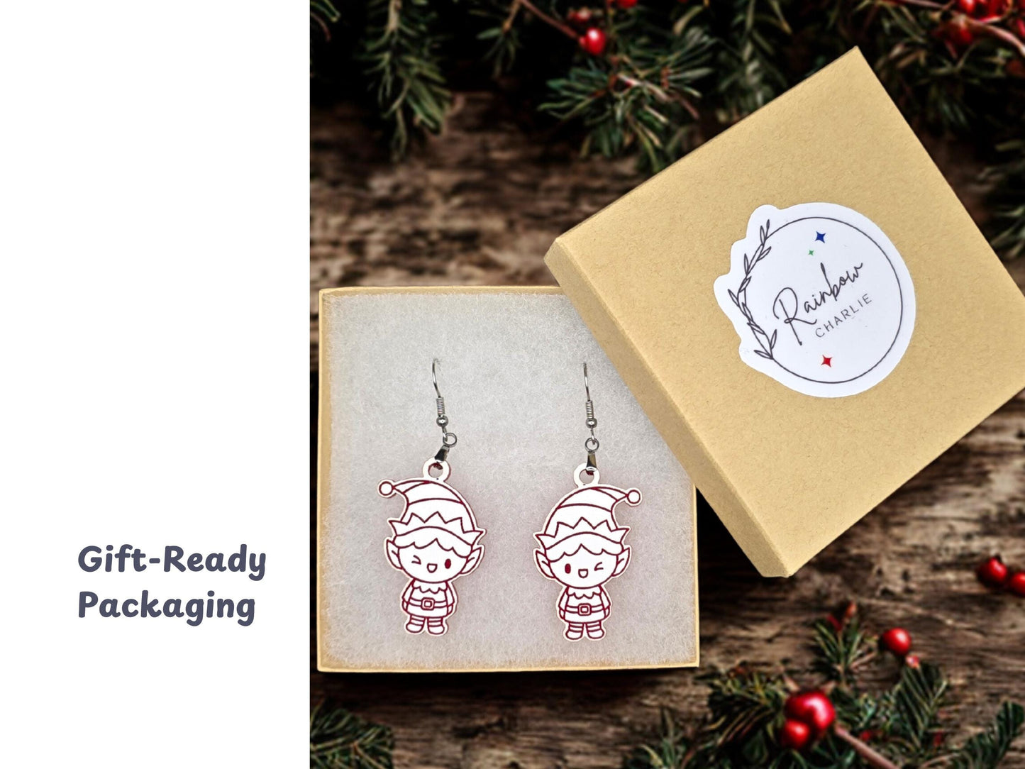 Elf earrings displayed inside a kraft jewelry gift box lined with soft white padding. The box lid, shown beside it, features a Rainbow Charlie logo sticker. Surrounding holiday greenery adds festive styling.