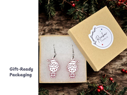 Elf earrings displayed inside a kraft jewelry gift box lined with soft white padding. The box lid, shown beside it, features a Rainbow Charlie logo sticker. Surrounding holiday greenery adds festive styling.