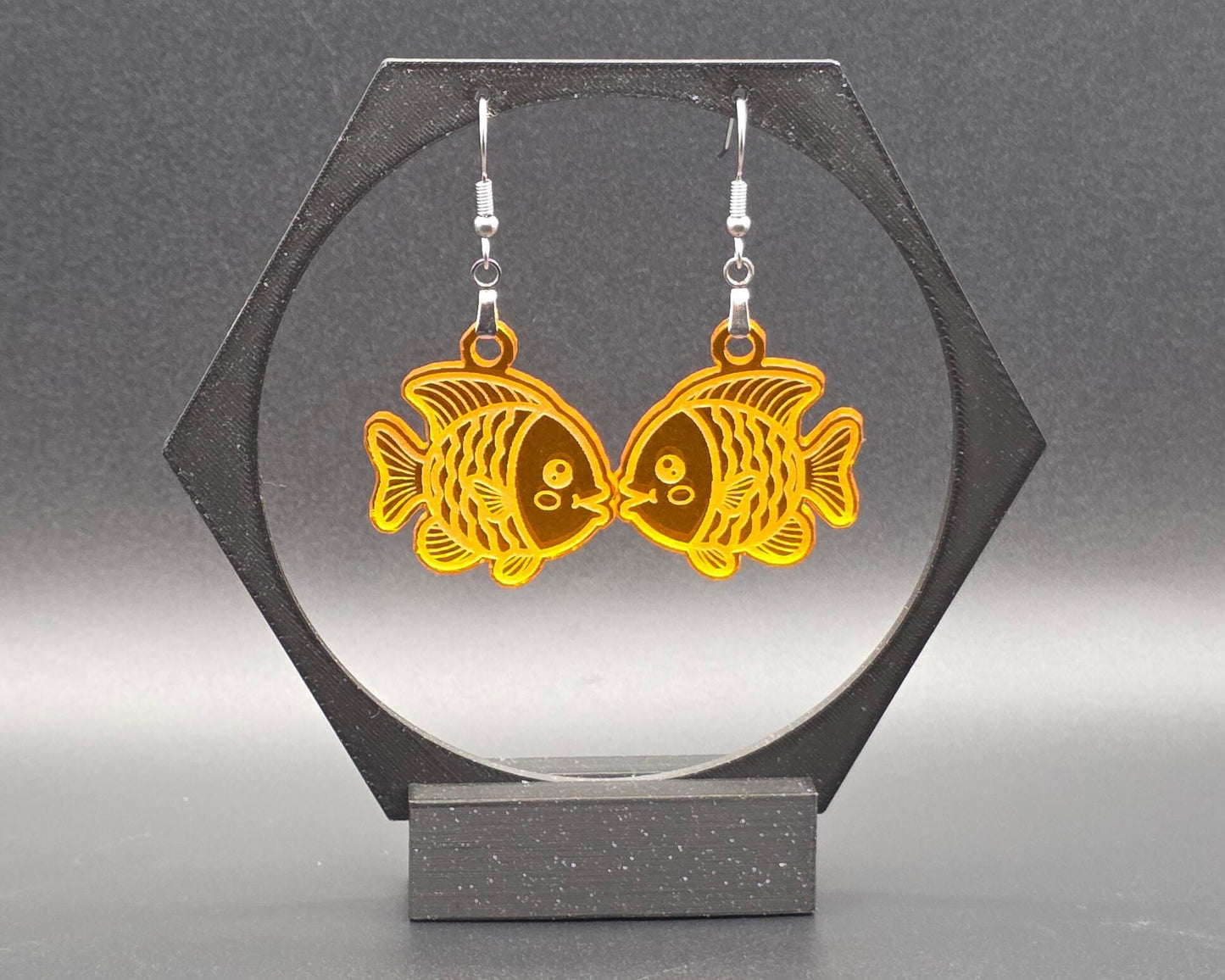 A pair of orange acrylic goldfish earrings with stainless steel hooks displayed on a hexagon stand against a gray background. The bright, laser-cut fish design adds a fun pop of color perfect for summer jewelry or beach accessories.