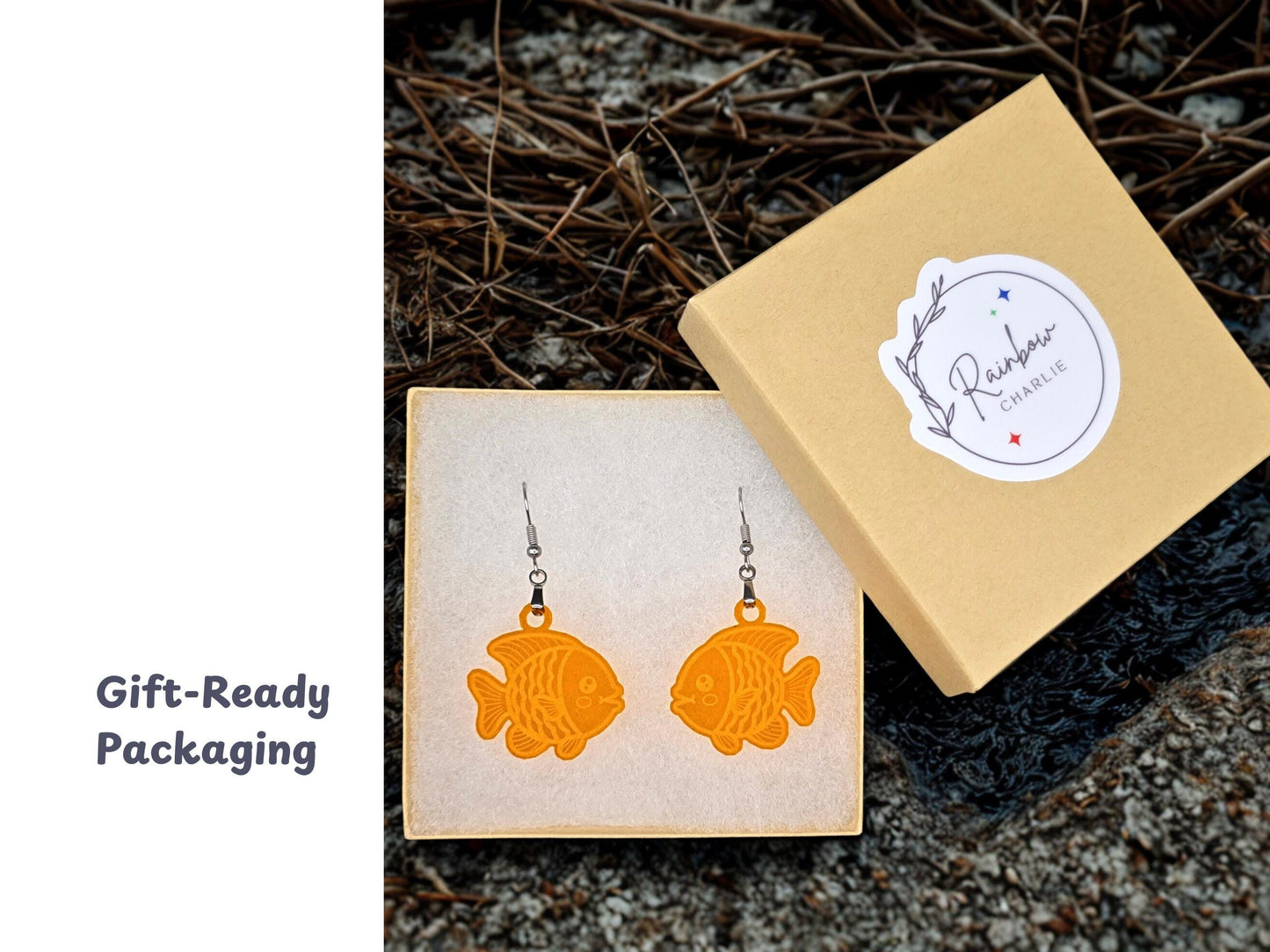 A pair of orange acrylic goldfish earrings displayed in a tan jewelry gift box with a Rainbow Charlie logo sticker. The box rests on textured natural ground, emphasizing the handmade, gift-ready presentation.