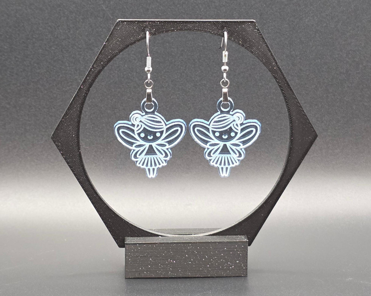 A pair of clear blue acrylic fairy earrings with engraved wing and dress details, hanging from nickel-free stainless steel hooks, displayed in a black hexagonal jewelry stand against a soft gray background. Handmade by Rainbow Charlie, whimsical and lightweight.