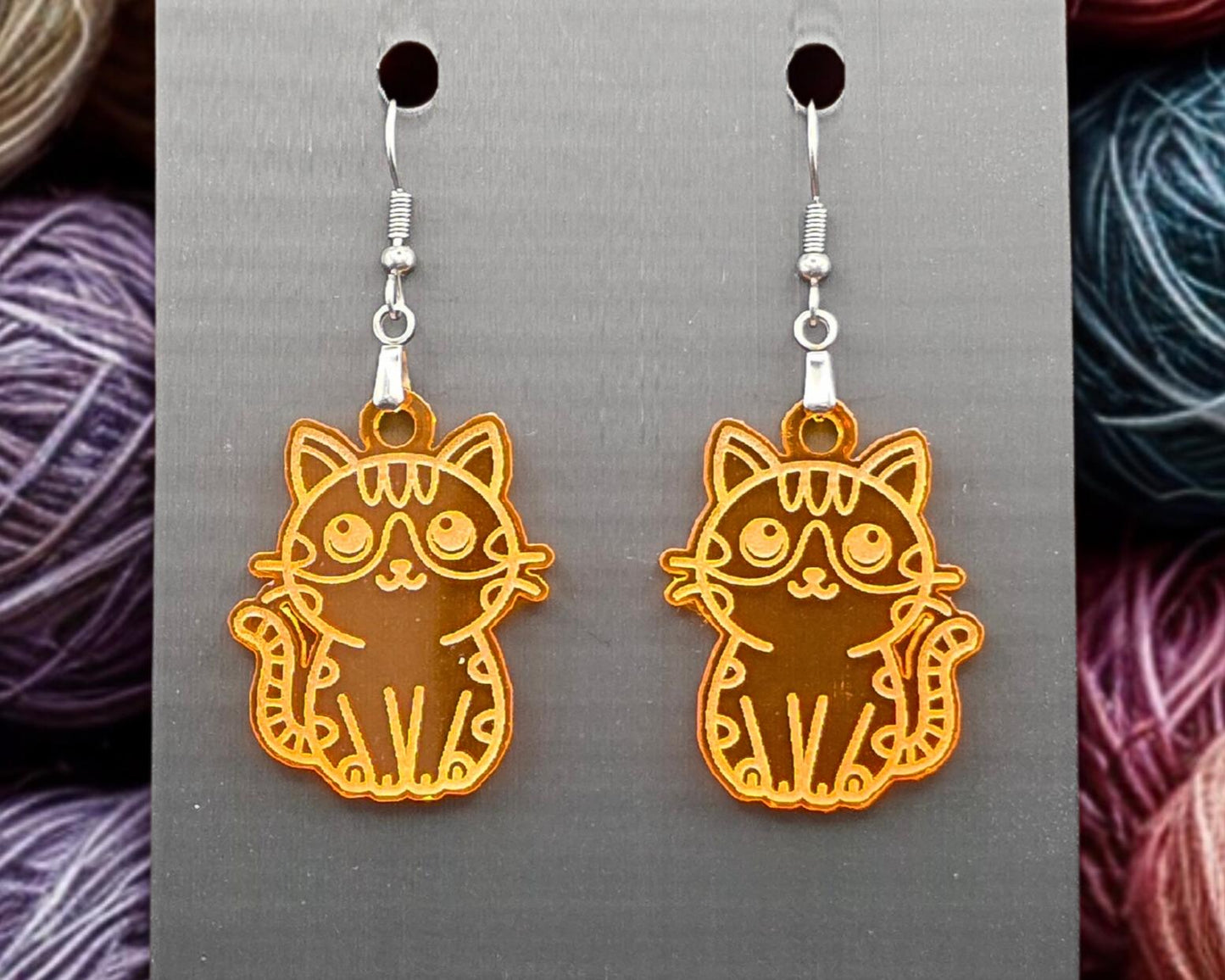 Orange acrylic cat earrings with reflective engraved details displayed on a gray backing card. Softly focused yarn balls appear along both edges of the image.