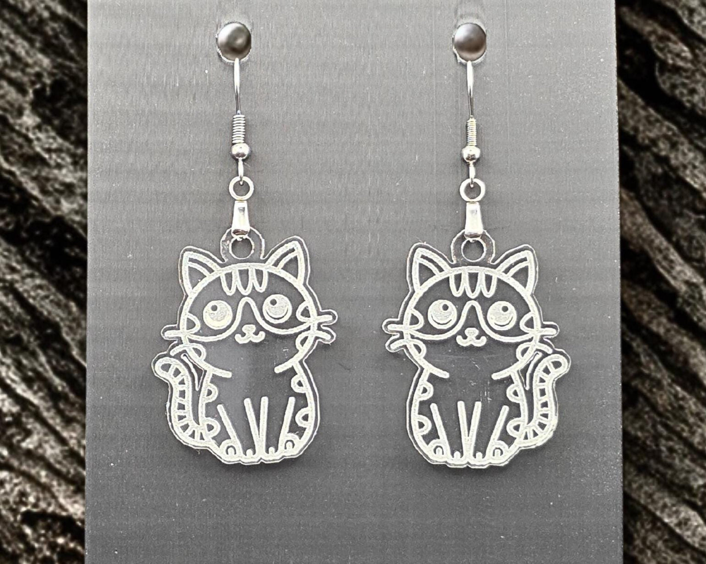 Clear acrylic cat earrings with engraved cat illustration, hanging on a gray display card. Stone-like gray granite texture borders both sides of the image.