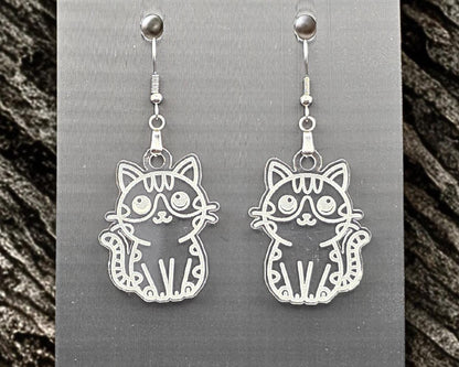 Clear acrylic cat earrings with engraved cat illustration, hanging on a gray display card. Stone-like gray granite texture borders both sides of the image.